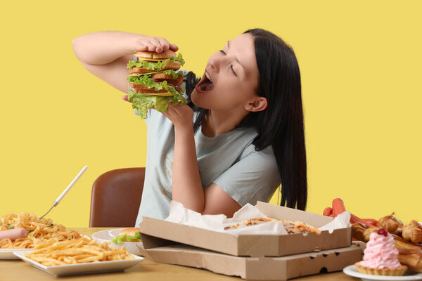 Молодая женщина с нездоровой пищей за столом на желтом фоне. Концепция переедания