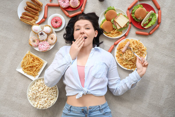 Удивлённая молодая женщина с нездоровой едой, лежащей на полу дома, вид сверху. Концепция переедания