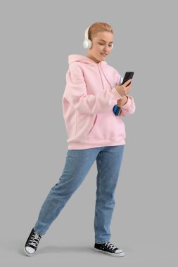 Kulaklık takan, kitapları olan, gri arka planda cep telefonu kullanan genç bir kadın.