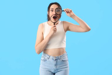Shocked young woman with magnifiers on blue background clipart