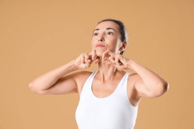 Olgun bir kadın bej arka planda kendine yüz masajı yapıyor.
