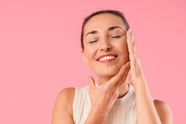 Güzel olgun bir kadın pembe arka planda kendine yüz masajı yapıyor.