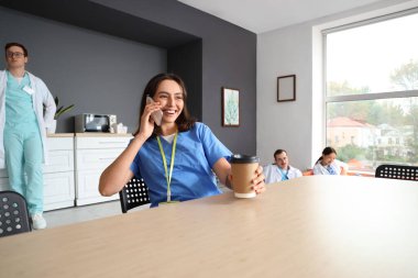 Elinde kahve fincanı olan kadın doktor hastane mutfağında cep telefonuyla konuşuyor.