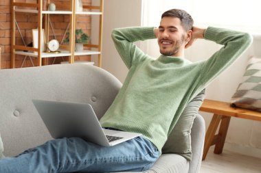 Evdeki kanepede dizüstü bilgisayarı olan yakışıklı genç adam.