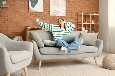 Evdeki kanepede dizüstü bilgisayarı olan genç ve güzel bir kadın.