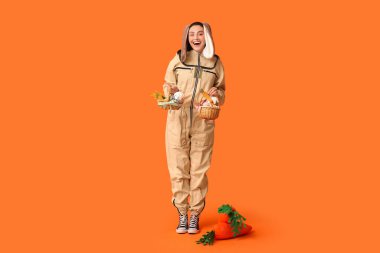 Paskalya sepetleri ve turuncu arka planda oyuncak havuçlarla tavşan kulaklı mutlu kadın arıcı.