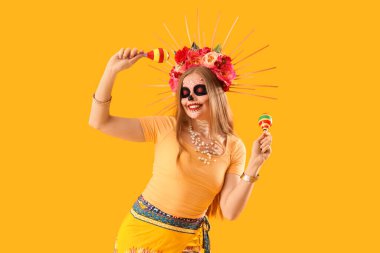 Yüzünde boyalı kafatası olan güzel genç bir kadın sarı arka planda marakas tutuyor. Meksika Ölüler Günü 'nün Kutlaması (El Dia de Muertos)