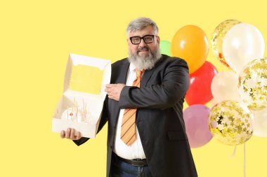 Doğum günü pastası ve sarı arka planda renkli balonları olan kıdemli iş adamı.