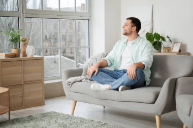 Evdeki gri koltukta oturan genç adam.