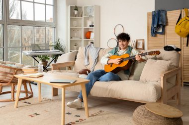 Erkek öğrenci evdeki kanepede gitar çalıyor.