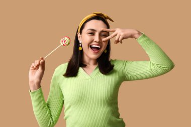 Tatlı lolipopu olan güzel genç bir kadın kahverengi arka planda zafer jesti gösteriyor.