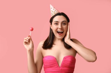 Pembe arka planda tatlı lolipopu olan parti şapkalı genç bir kadın.