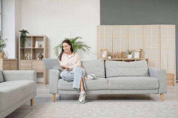 Красивая молодая женщина с ноутбуком сидит на сером диване в гостиной