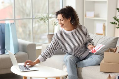 Genç Afro-Amerikalı kadın, hediye kartı ve laptop ile evdeki kanepede alışveriş yapıyor.