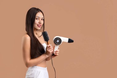 Beautiful young happy Asian woman with hairdryer and brush on brown background clipart