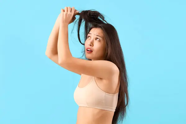 Beautiful young stressed Asian woman trying to brush tangled hair on blue background
