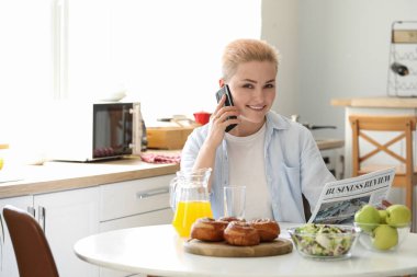 Gazeteli genç bir kadın mutfakta cep telefonuyla konuşuyor.