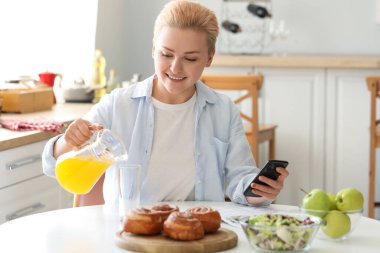 Cep telefonu olan genç bir kadın mutfakta cama meyve suyu döküyor.