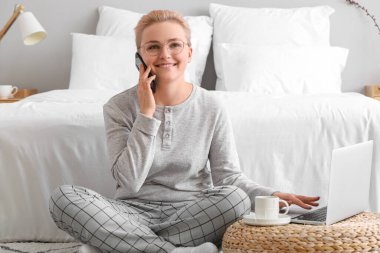 Yatak odasında dizüstü bilgisayarı olan genç bir kadın cep telefonuyla konuşuyor.