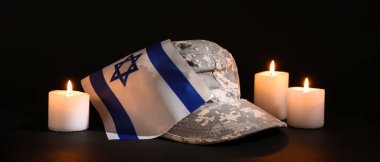 İsrail bayrağı taşıyan askeri şapka ve koyu arkaplanda yanan mumlar.