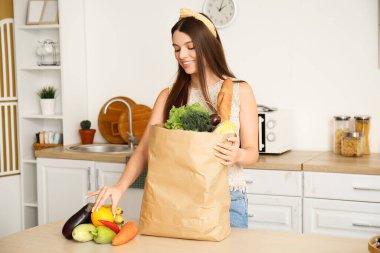 Mutfaktaki masada elinde bir torba taze sebze ve meyve olan genç bir kadın.