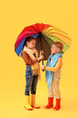 Sarı arka planda şemsiyesi olan lastik çizmeli küçük çocuklar.