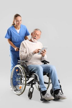 Tekerlekli sandalyedeki yaşlı adam tablet bilgisayar kullanıyor ve arka planda hemşire var.