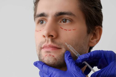 Genç adamın yüzünde şırınga ve işaretler olan estetik cerrah, yakın plan.