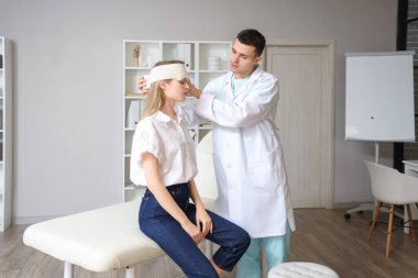 Erkek doktor klinikte beyin sarsıntısı geçiren kadının kafasını bandajlıyor.