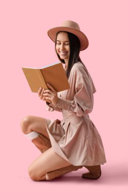 Şapkalı genç bir kadın pembe arka plan kitabı okuyor.