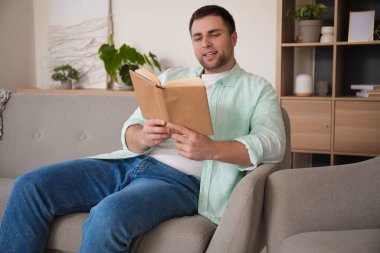 Genç adam evdeki gri koltukta kitap okuyor.
