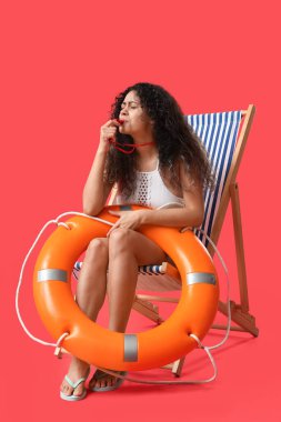 Yüzük şamandıralı güzel Afro-Amerikalı kadın cankurtaran kırmızı arka planda güvertede oturuyor.