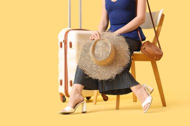 Sarı arka planda oturan, bavulu ve yaz şapkası olan bir bayan yolcu.