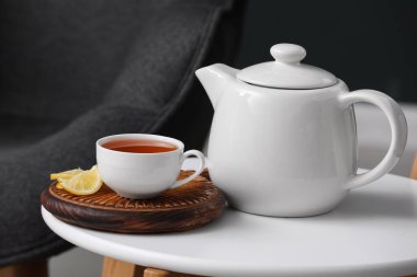 Bir fincan sıcak demlenmiş çay, bir dilim limon ve demlik siyah bulanık arka planda.