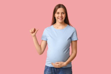 Güzel, genç, hamile bir kadın pembe arka plandaki zarif mavi tişörtü işaret ediyor.