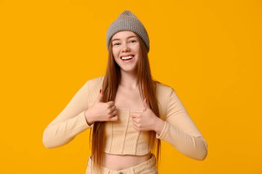 Üstte havalı genç bir kız ve sarı arka planda baş parmağını gösteren örgü bir şapka.