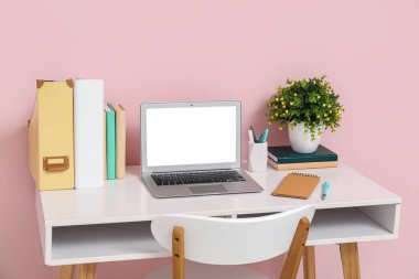 Modern dizüstü bilgisayarı, ev bitkisi ve pembe duvara yakın kitapları olan şık bir iş yeri.