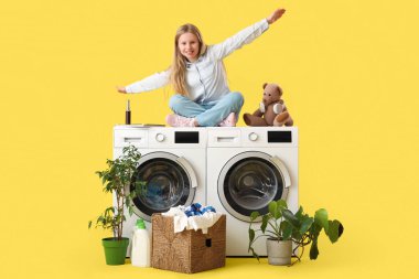 Oyuncak ayılı bir kız, saksı ve sepetin yanındaki çamaşır makinesinde oturuyor. Çamaşırları sarı arka planda.
