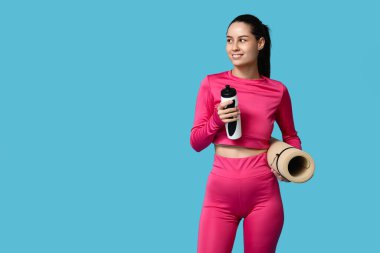 Mavi arka planda su şişesi ve yoga minderi olan genç ve sportif bir kadın.