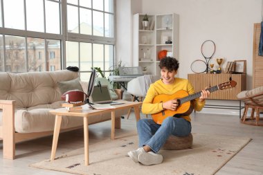 Erkek öğrenci evde dolaşırken gitar çalıyor.