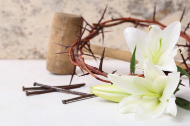 Composition crown of thorns, lilies, nails and hammer on white table near light wall clipart