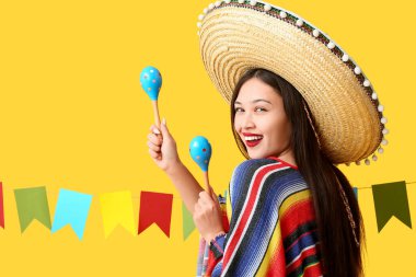 Meksika şapkalı ve pançolu mutlu genç kadın sarı arka planda marakas tutuyor.