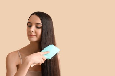 Güzel genç bir kadın bej arka planda uzun kahverengi düz saçlarını tarıyor.