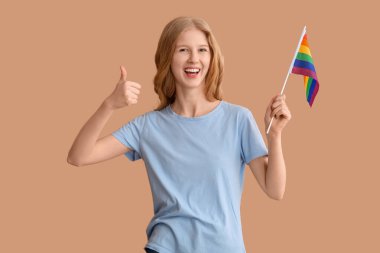 LGBT bayrağıyla güzel, genç, mutlu bir kadın bej arka planda baş parmak hareketi gösteriyor.