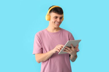 Kulaklıklı, tabletli yakışıklı genç adam mavi arka planda sesli kitap dinliyor.