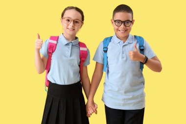 Sırt çantalı mutlu küçük öğrenciler sarı arka planda baş parmak hareketi gösteriyorlar.