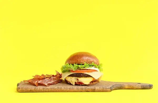 Board with tasty burger and bacon on yellow background
