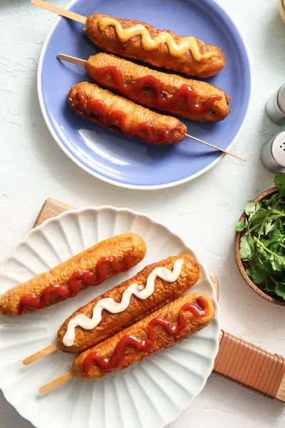 Plates of tasty corn dogs with different sauces on white background