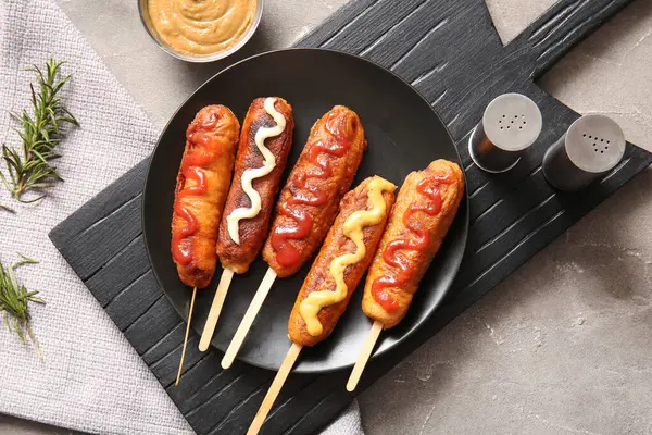 Plate of tasty corn dogs with different sauces on grey background