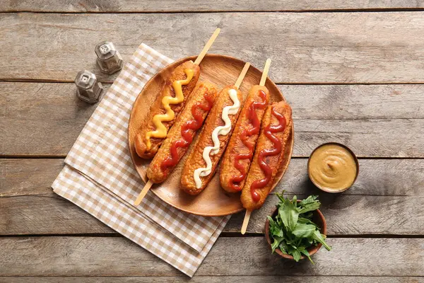 Plate of tasty corn dogs with different sauces on wooden background